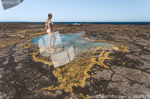 Image of Woman stands by a small rock pool edged with yellow seaweed