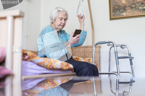 Image of Elderly 96 years old woman reading phone message while sitting on medical bed supporting her by holder.