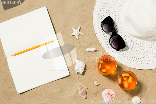 Image of notebook, cocktails, hat and shades on beach sand