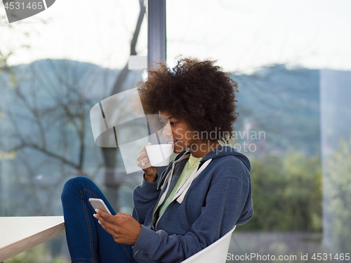 Image of black woman drinking coffee and using a mobile phone  at home