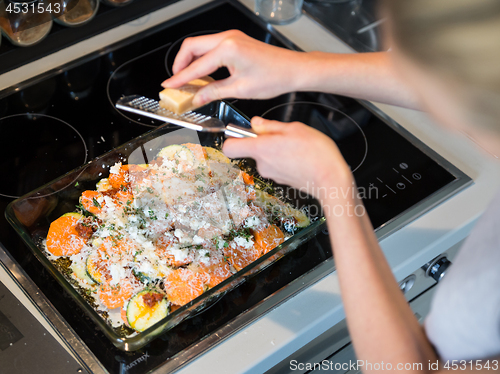 Image of Female chef grinding parmesan cheese on row vegetarian dish ingredients in glass baking try before placing it into oven. Healthy home-cooked everyday vegetarian food