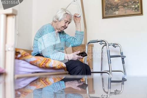 Image of Elderly 96 years old woman reading phone message while sitting on medical bed supporting her by holder.