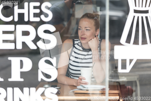 Image of Young caucasian woman sitting alone in coffee shop thoughtfully leaning on her hand, looking trough the window