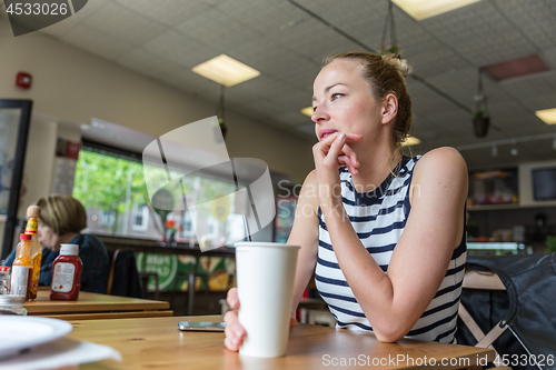 Image of Young caucasian woman sitting alone in coffee shop thoughtfully leaning on her hand, looking trough the window