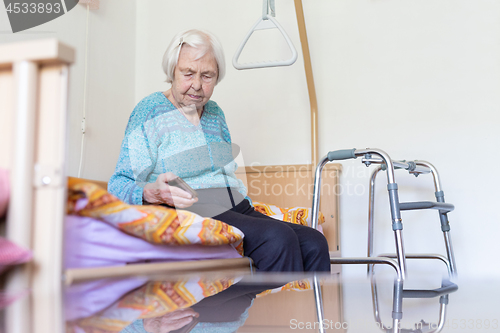 Image of Elderly 96 years old woman reading phone message while sitting on medical bed supporting her by holder.