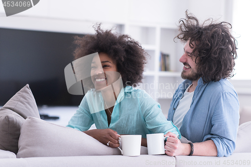 Image of multiethnic couple sitting on sofa at home drinking coffe