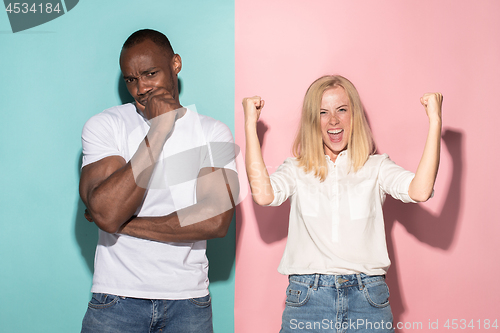 Image of Closeup portrait of young couple, man, woman. One being excited happy smiling, other serious, concerned, unhappy on pink and blue background. Emotion contrasts