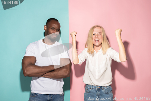 Image of Closeup portrait of young couple, man, woman. One being excited happy smiling, other serious, concerned, unhappy on pink and blue background. Emotion contrasts