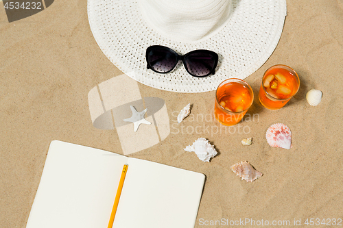 Image of notebook, cocktails, hat and shades on beach sand