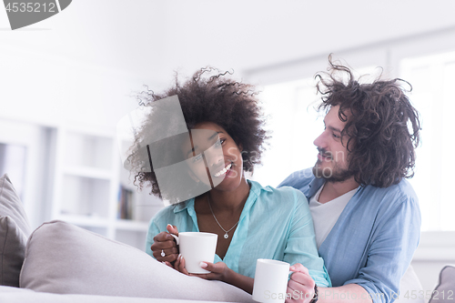 Image of multiethnic couple sitting on sofa at home drinking coffe