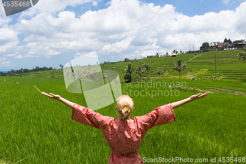 Image of Relaxed fashionable caucasian woman wearing red asian style kimono, arms rised to sky, enjoying pure nature at beautiful green rice fields on Bali island