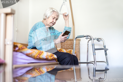 Image of Elderly 96 years old woman reading phone message while sitting on medical bed supporting her by holder.