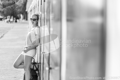 Image of Blonde young female traveler wearing summer style clothing, leaning against retro blue beach dressing rooms at summer time vacation in Sistiana, Italy