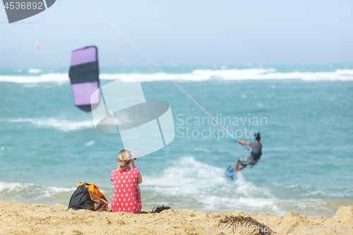 Image of Girl sitting on sandy beach watching kite surfer in the sea.