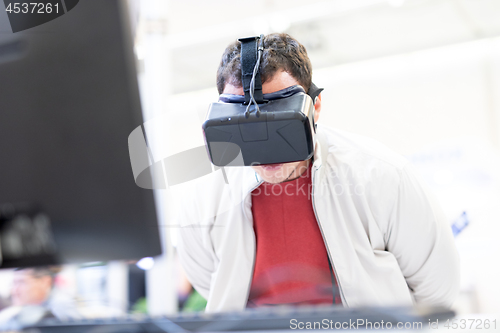 Image of Young man wearing virtual reality headset and gesturing while sitting at his desk in creative office