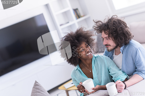 Image of multiethnic couple sitting on sofa at home drinking coffe