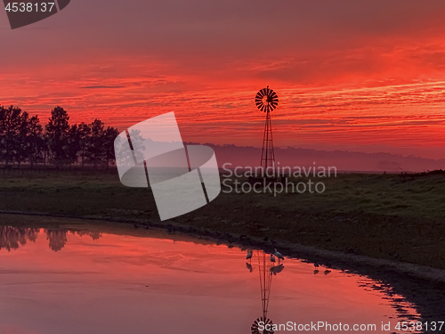 Image of Light morning fog, windmill, pond with red sunrise sky in rural 