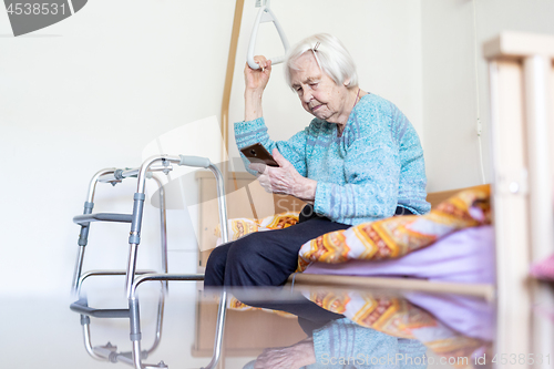 Image of Elderly 96 years old woman reading phone message while sitting on medical bed supporting her by holder.