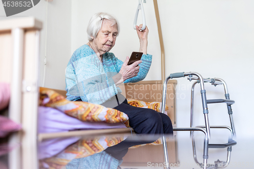 Image of Elderly 96 years old woman reading phone message while sitting on medical bed supporting her by holder.