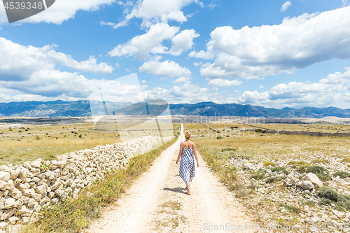 Image of Caucasian young woman in summer dress holding bouquet of lavender flowers while walking outdoor through dry rocky Mediterranean Croatian coast lanscape on Pag island in summertime