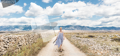Image of Caucasian young woman in summer dress holding bouquet of lavender flowers while walking outdoor through dry rocky Mediterranean Croatian coast lanscape on Pag island in summertime