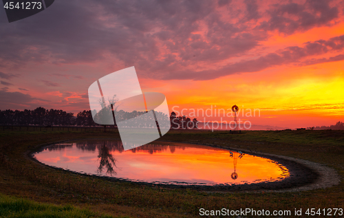 Image of Windmill by pond in rural field with vivid sunrise