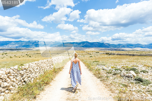 Image of Caucasian young woman in summer dress holding bouquet of lavender flowers while walking outdoor through dry rocky Mediterranean Croatian coast lanscape on Pag island in summertime