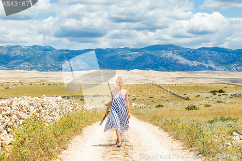 Image of Caucasian young woman in summer dress holding bouquet of lavender flowers while walking outdoor through dry rocky Mediterranean Croatian coast lanscape on Pag island in summertime