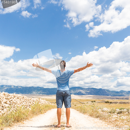 Image of Rear view of casual sporty man standing on a dirt country road rising hands up to the clouds on a blue summer sky. Freedom and travel adventure concept.