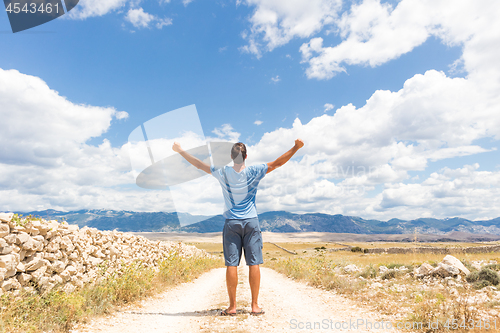 Image of Rear view of casual sporty man standing on a dirt country road rising hands up to the clouds on a blue summer sky. Freedom and travel adventure concept.