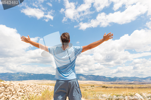 Image of Rear view of casual sporty man standing on a dirt country road rising hands up to the clouds on a blue summer sky. Freedom and travel adventure concept.