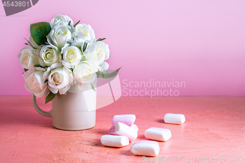 Image of Small white and pink marshmallows are scattered on a pale pink background next to a vase of roses. Place for text.