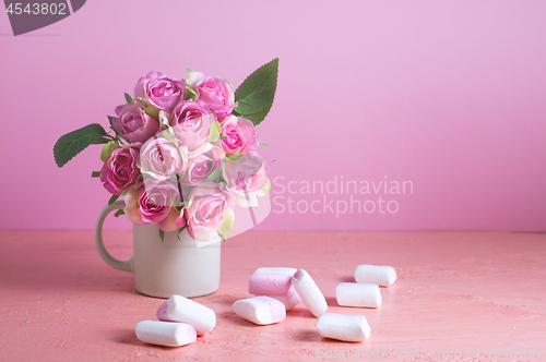 Image of Small white and pink marshmallows are scattered on a pale pink background next to a vase of roses. Place for text.