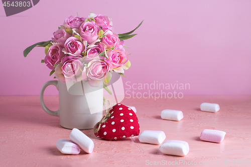 Image of Small white and pink marshmallows are scattered on a pale pink background next to a vase of roses. Place for text.