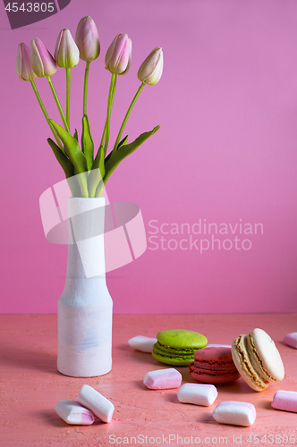 Image of Macaroons and small white and pink marshmallows are scattered on a pale pink background next to a vase of tulips. Place for text.