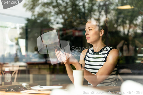 Image of Thoughtful caucasian woman holding mobile phone while looking through the coffee shop window during coffee break.