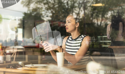 Image of Thoughtful caucasian woman holding mobile phone while looking through the coffee shop window during coffee break.