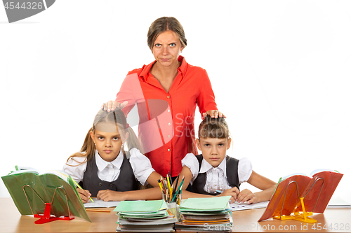 Image of Angry teacher stands behind students sitting at her desk and puts her hands on the heads of children