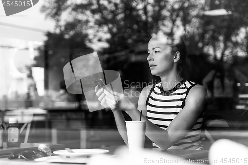 Image of Thoughtful caucasian woman holding mobile phone while looking through the coffee shop window during coffee break.