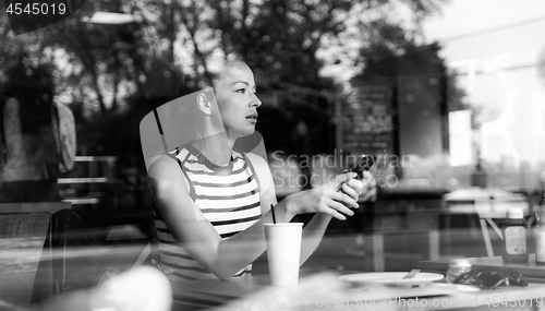 Image of Thoughtful caucasian woman holding mobile phone while looking through the coffee shop window during coffee break.