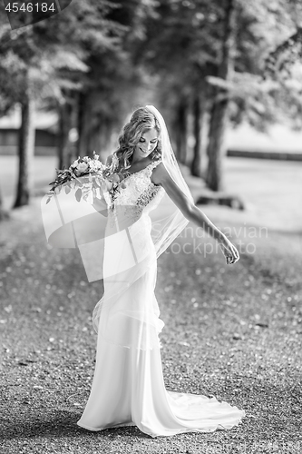 Image of Full length portrait of beautiful sensual young blond bride in long white wedding dress and veil, holding bouquet outdoors in natural background.