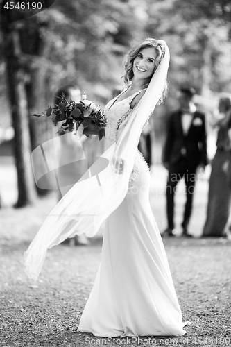 Image of Full length portrait of beautiful sensual young blond bride in long white wedding dress and veil, holding bouquet outdoors in natural background.