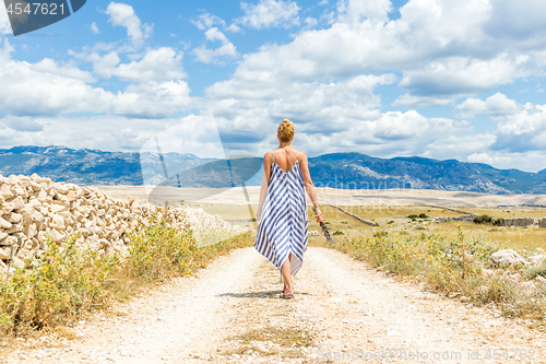Image of Rear view of woman in summer dress holding bouquet of lavender flowers while walking outdoor through dry rocky Mediterranean Croatian coast lanscape on Pag island in summertime