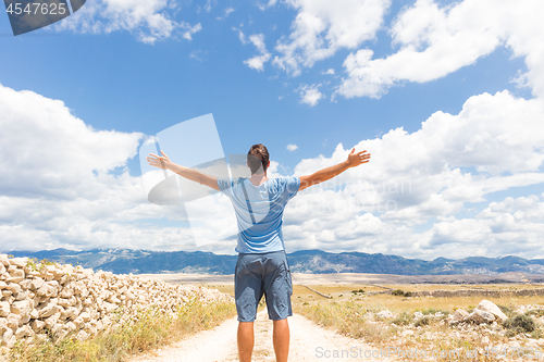 Image of Rear view of casual sporty man standing on a dirt country road rising hands up to the clouds on a blue summer sky. Freedom and travel adventure concept.