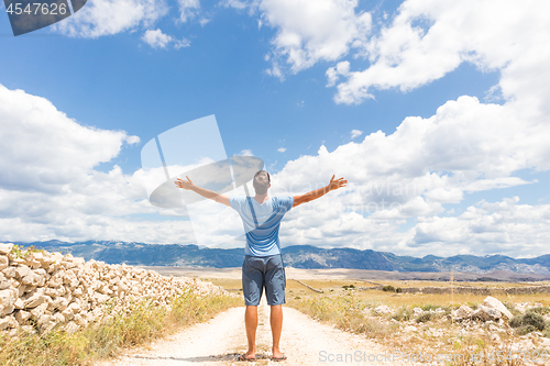 Image of Rear view of casual sporty man standing on a dirt country road rising hands up to the clouds on a blue summer sky. Freedom and travel adventure concept.