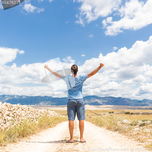 Image of Rear view of casual sporty man standing on a dirt country road rising hands up to the clouds on a blue summer sky. Freedom and travel adventure concept.