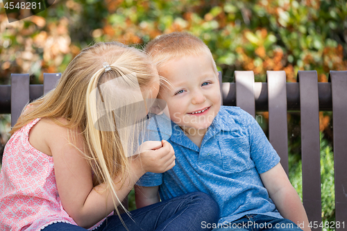 Image of Young Sister and Brother Whispering Secrets On The Bench At The 