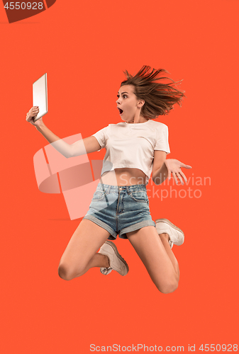 Image of Image of young woman over red background using laptop computer or tablet gadget while jumping.