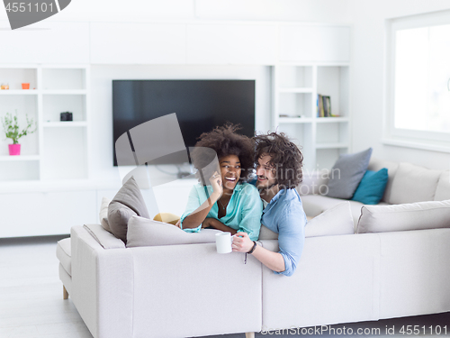Image of multiethnic couple sitting on sofa at home drinking coffe