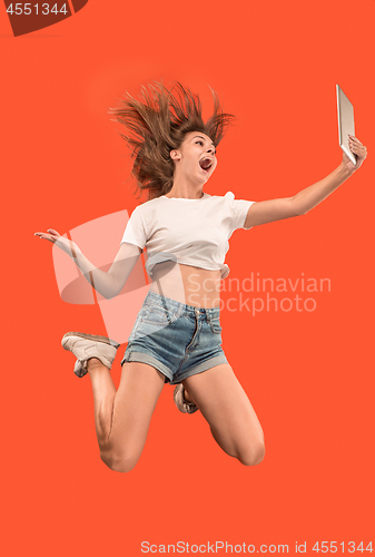 Image of Image of young woman over red background using laptop computer or tablet gadget while jumping.